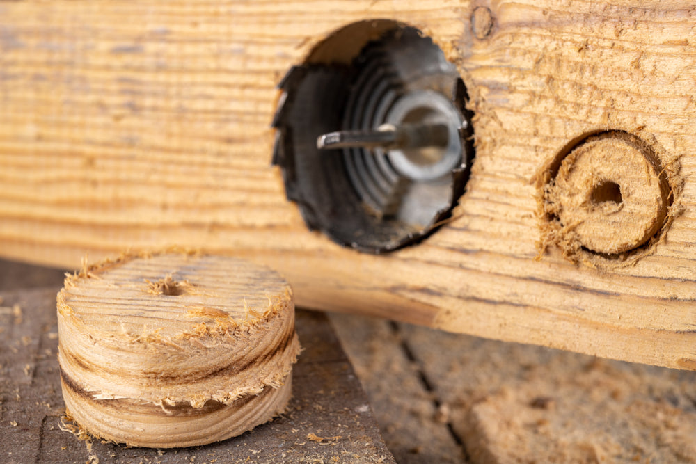 A close-up of a piece of wood with a circular hole cut by a hole saw drill bit, with the removed wooden disc lying nearby.