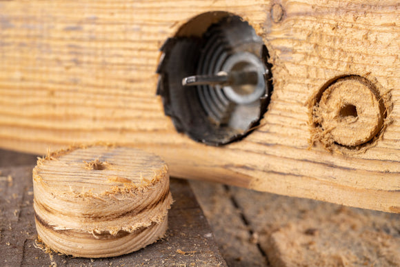 A close-up of a piece of wood with a circular hole cut by a hole saw drill bit, with the removed wooden disc lying nearby.